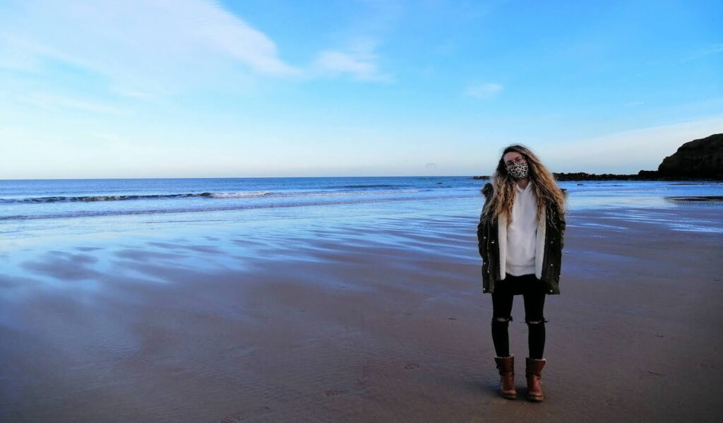 Holly standing on a beach with a face mask and big coat on, with the sea behind her and blue sky above her