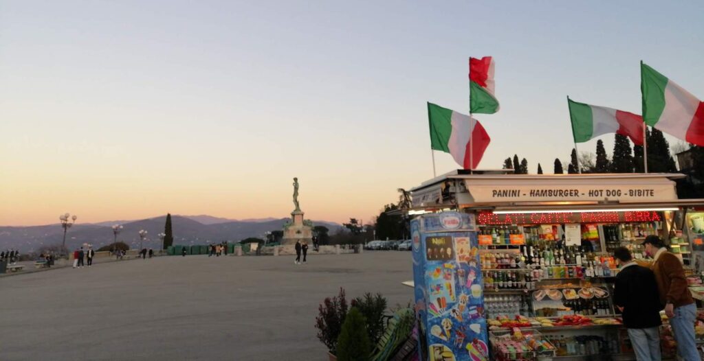 On the right, a kiosk with Italian flags on top blowing in the breeze. To the left, Piazzale Michelangelo and a copy of the David in the background against the hills of Florence.